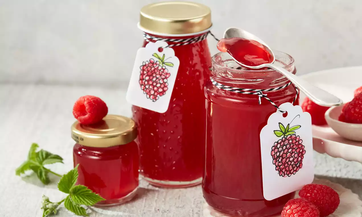 Glänzendes rotes Himbeergelee in Gläsern mit goldenen Deckeln, dekoriert mit Etiketten, umgeben von frischen Himbeeren und grünen Blättern