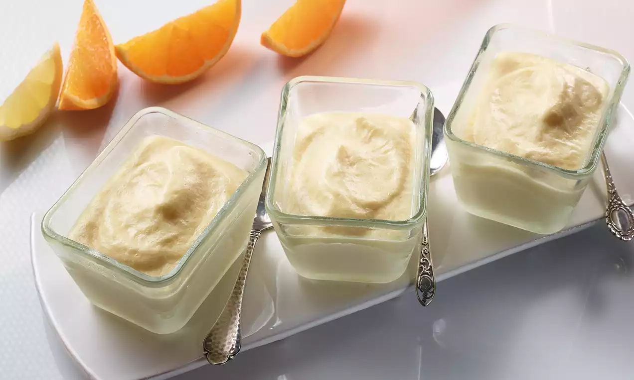 Crèmes citron pâle à la texture lisse servies dans des verres carrés sur un plateau blanc avec des tranches d'orange en arrière-plan