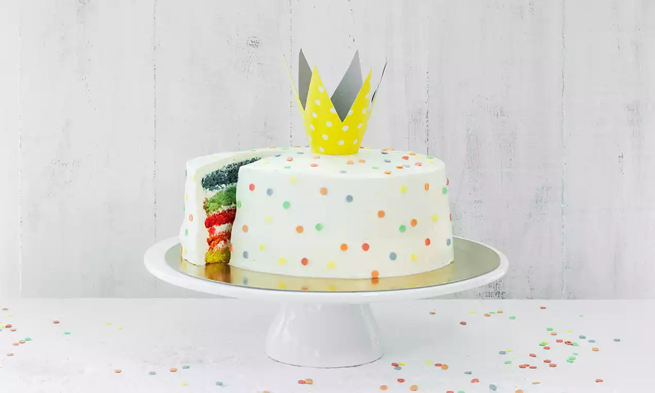 Witte ronde taart met kleurige stipjes en een gele kroon, regenbooglagen zichtbaar bij een plak, op een wit bord met confetti