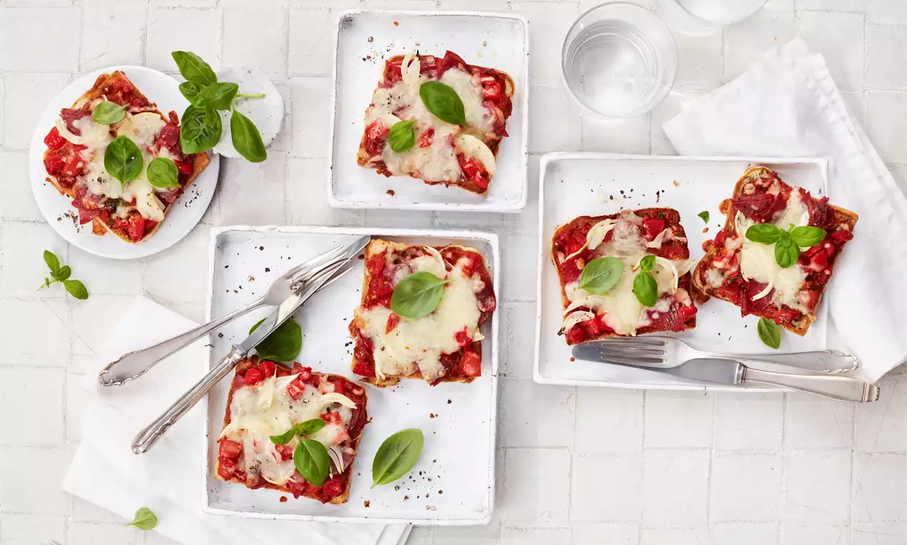 Pizza tost pravokutnog oblika s hrskavom podlogom, zapečenom sirom, rajčicom i svježim listovima bosiljka na bijelim tanjirima