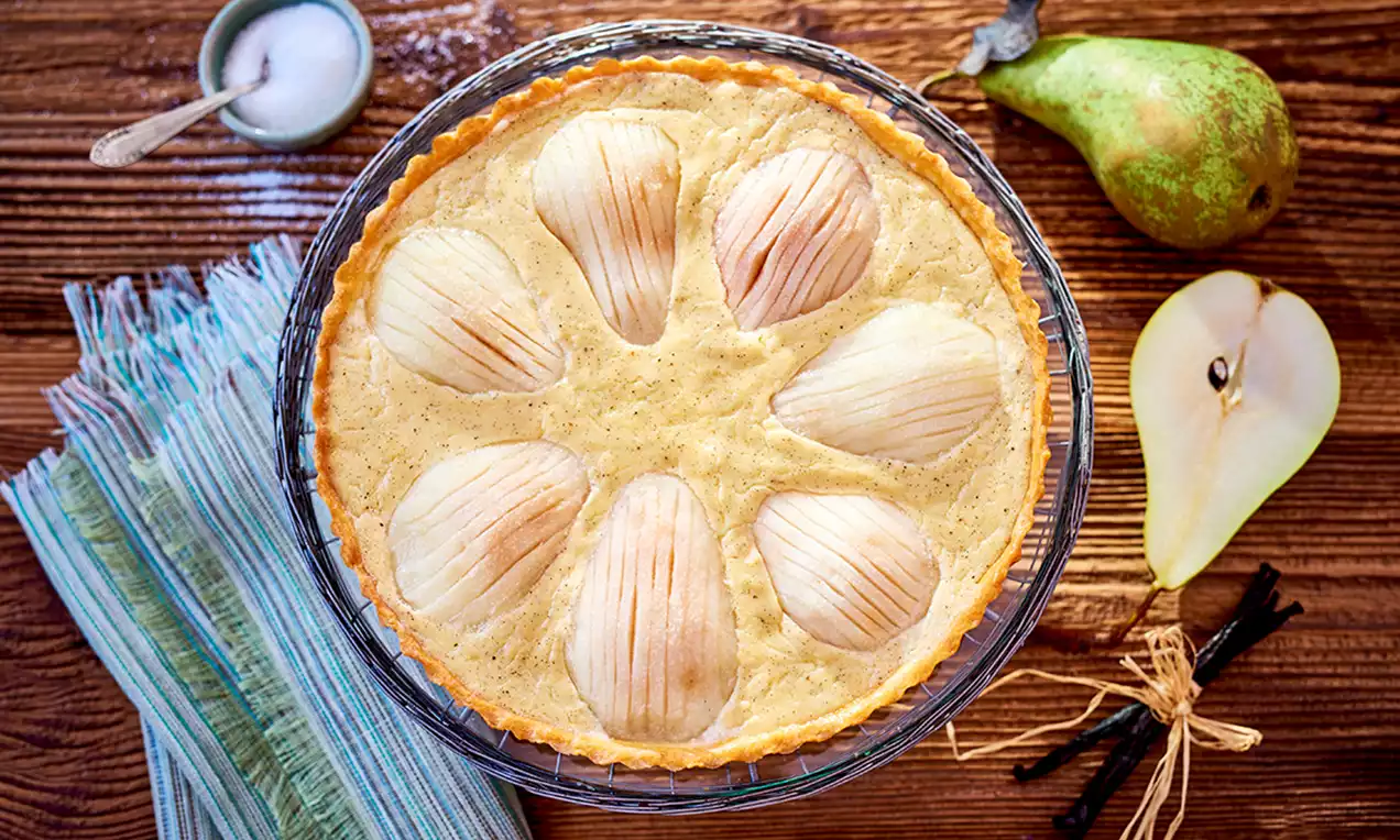 Tart s valovitim polovicama krušaka u zlatnoj vanil kremi na tankoj smeđoj podlozi, poslužen na drvenoj podlozi s kruškom i vanilom