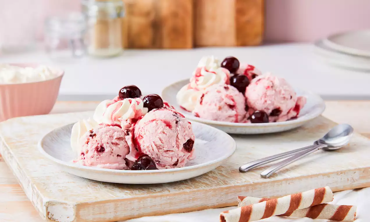 Na bijelom tanjuru kuglice svijetloružičastog sladoleda s tamnim Amarena višnjama, ukrašene hrpicama šlaga i preljevom od višnje