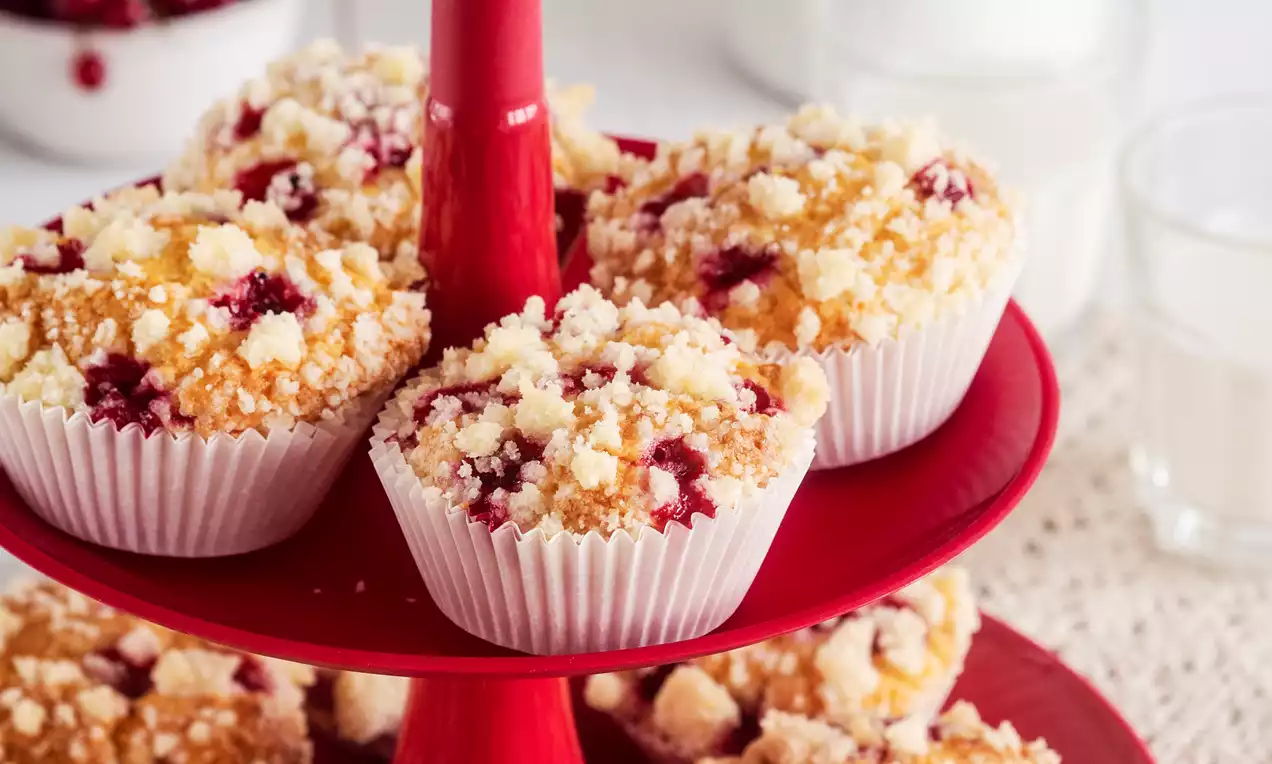 Kruche babeczki jogurtowe z kawałkami czerwonych porzeczek i złocistą kruszonką, w białych papilotkach, ułożone na czerwonym piętrowym talerzu