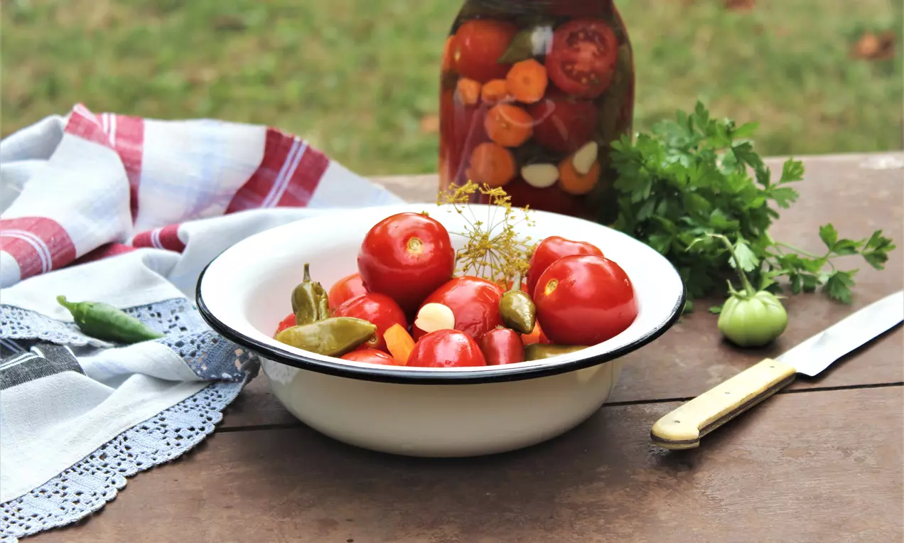 Червени домати с гладка кожа, подредени в бяла емайлирана купа със зелени пиперки, чесън и копър, фон с буркан и свежи билки