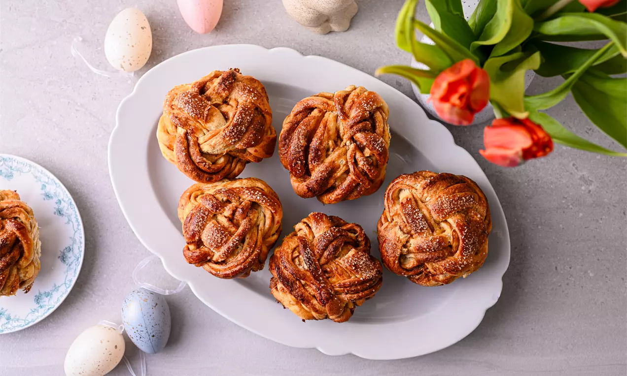 Козуначени мъфини с кардамон и портокал с извита форма и златиста коричка са подредени върху бяла плоска чиния до цветя и шарени яйца