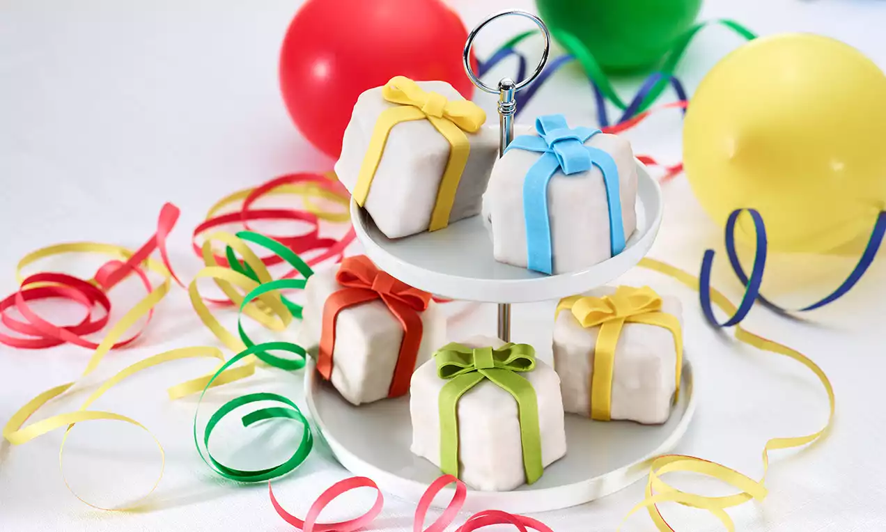 Dolcetti cubici ricoperti di glassa bianca con nastri colorati di zucchero disposti su un'alzata a due piani, circondati da festoni e palloncini