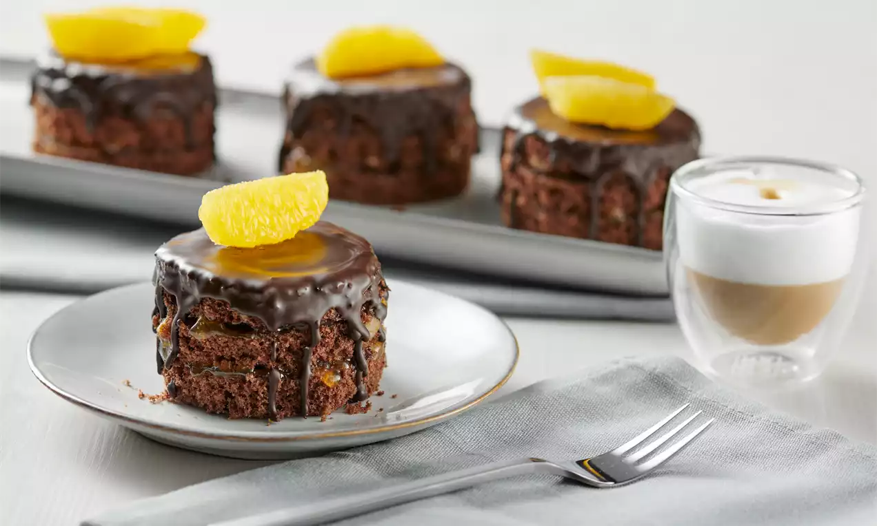 Kleine runde Törtchen aus dunklem Schokoteig mit glänzender Schokoglasur, garniert mit orange-gelben Orangenstückchen auf weißem Teller