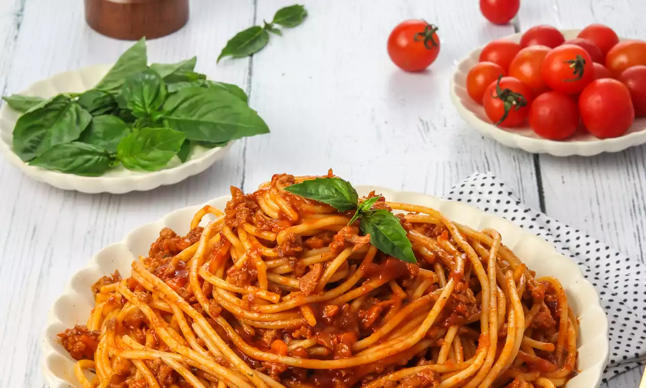 Spaghetti strands coated in rich red Bolognese sauce with crumbled meat, garnished with vibrant basil leaves on a white scalloped plate