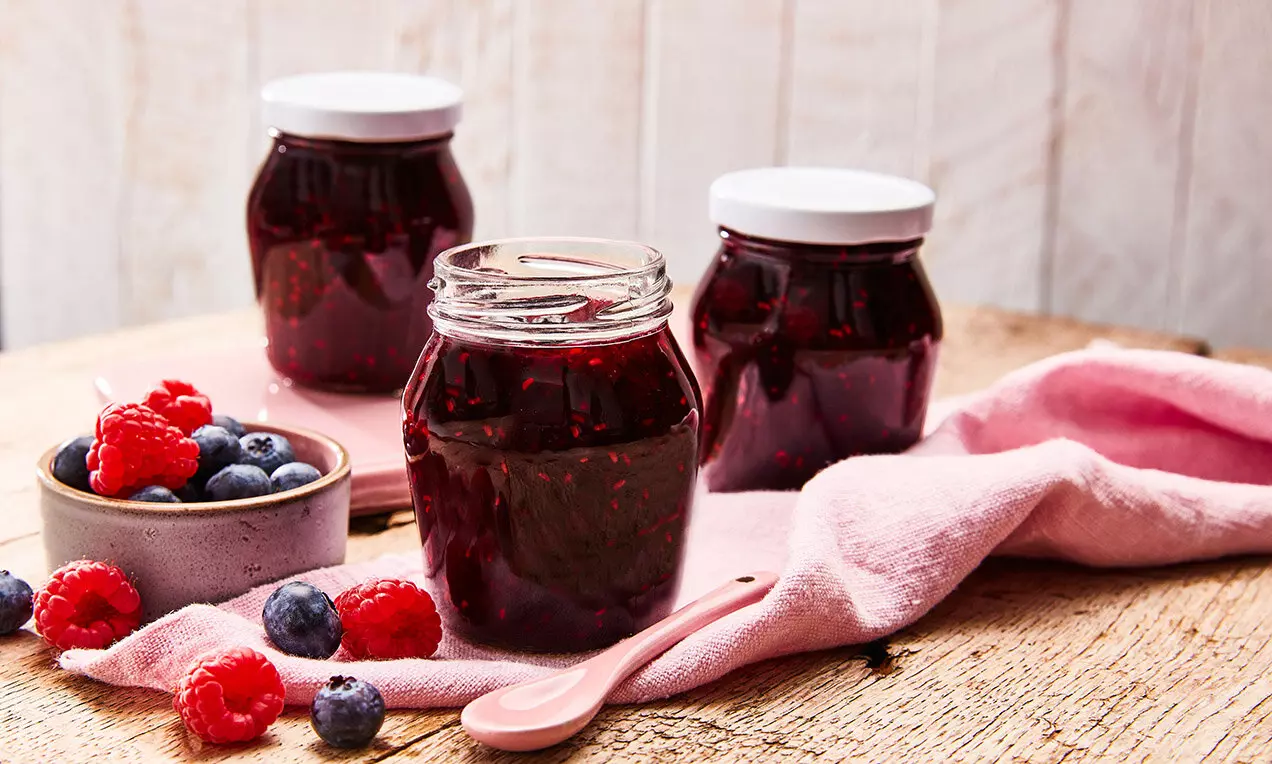 Тъмночервени компакти бурканчета със сладко из прозрачна стъклена текстура, заобиколени от свежи малини и боровинки върху дървена маса