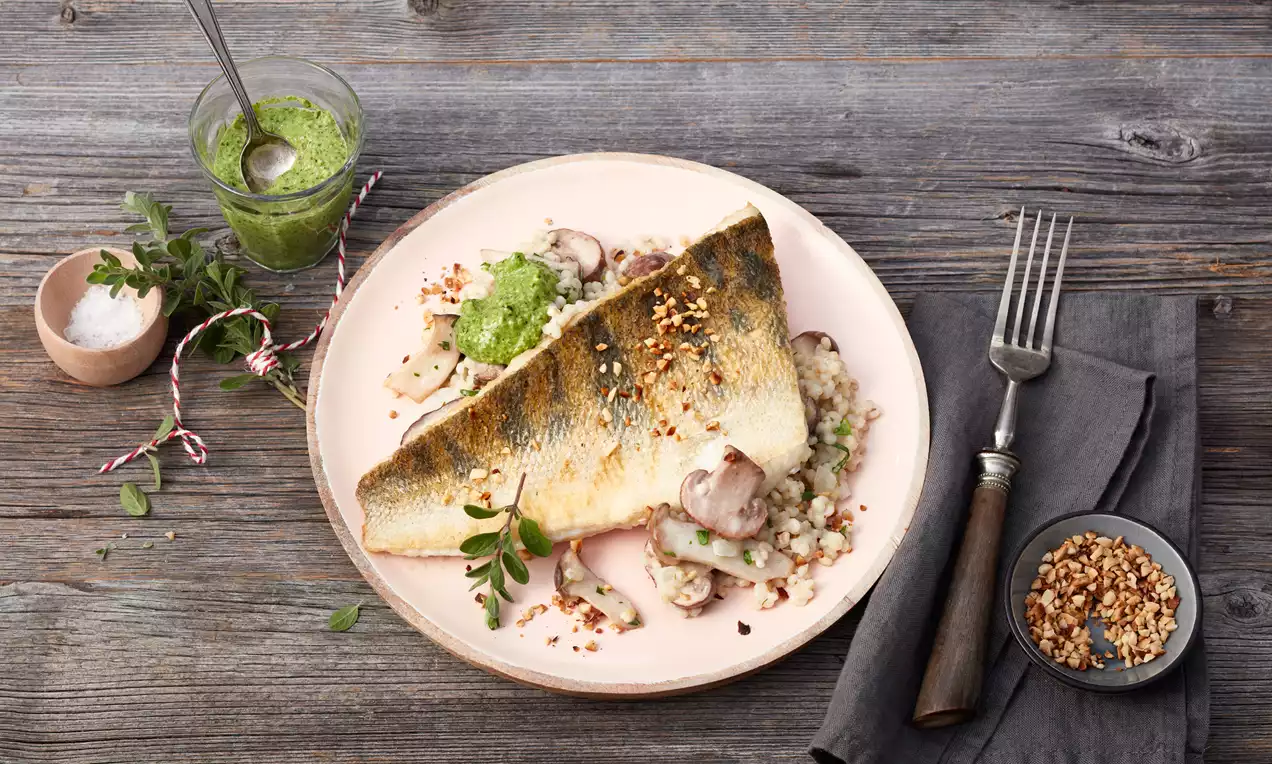 Goldbraun gebratener Zander auf cremigem Pilzrisotto mit grünem Feldsalat-Pesto, angerichtet auf einem hellen Teller mit Holzuntergrund