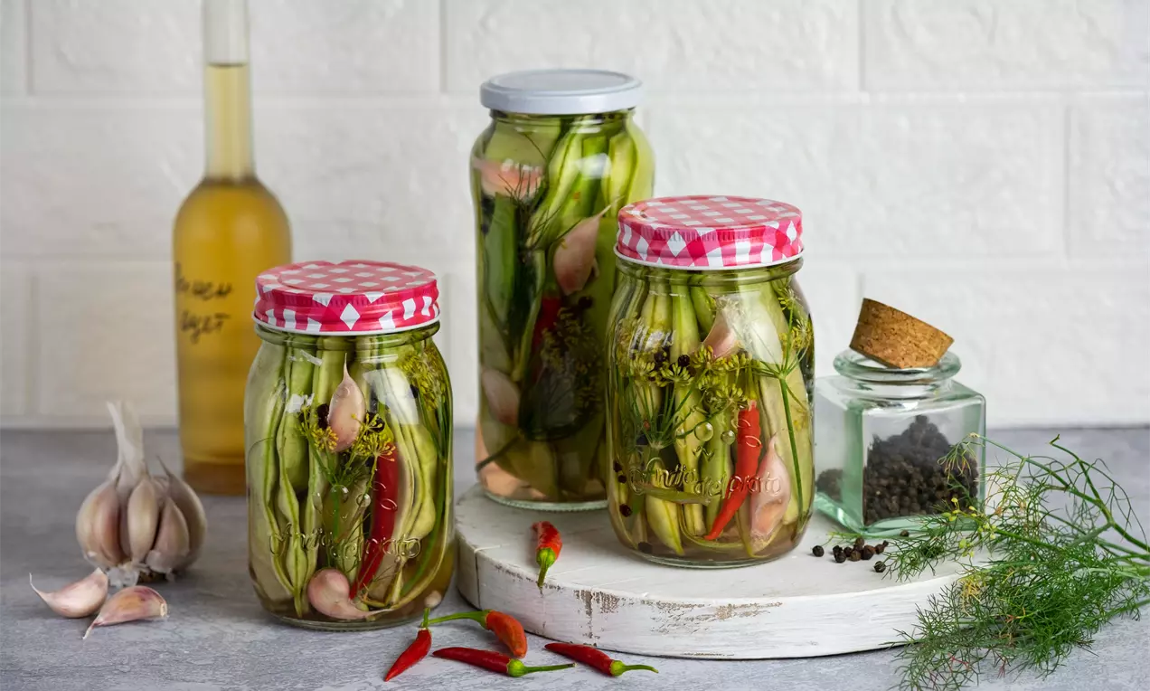 Тънки, зелени стръкове фасул с яркочервени чушки и цели скилидки чесън в буркани с прозрачен стъклен корпус и капаци с шарка на червени карета