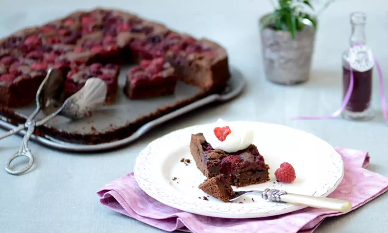 Chocoladebrownie met donkerbruine korst en sappige frambozen, geserveerd met slagroom en een hele framboos op een wit bord met roze servet