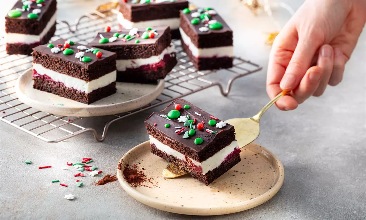 Шоколадов сладкиш със слой бял крем, декориран с червени и зелени захарни топчета, поднесен върху светла керамична чиния