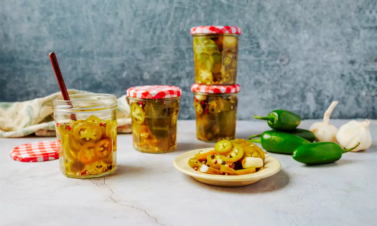Тънки зелени кръгчета от халапеньо в прозрачен буркан с червена капачка върху светла повърхност, сервирани на чиния до пресни люти чушки
