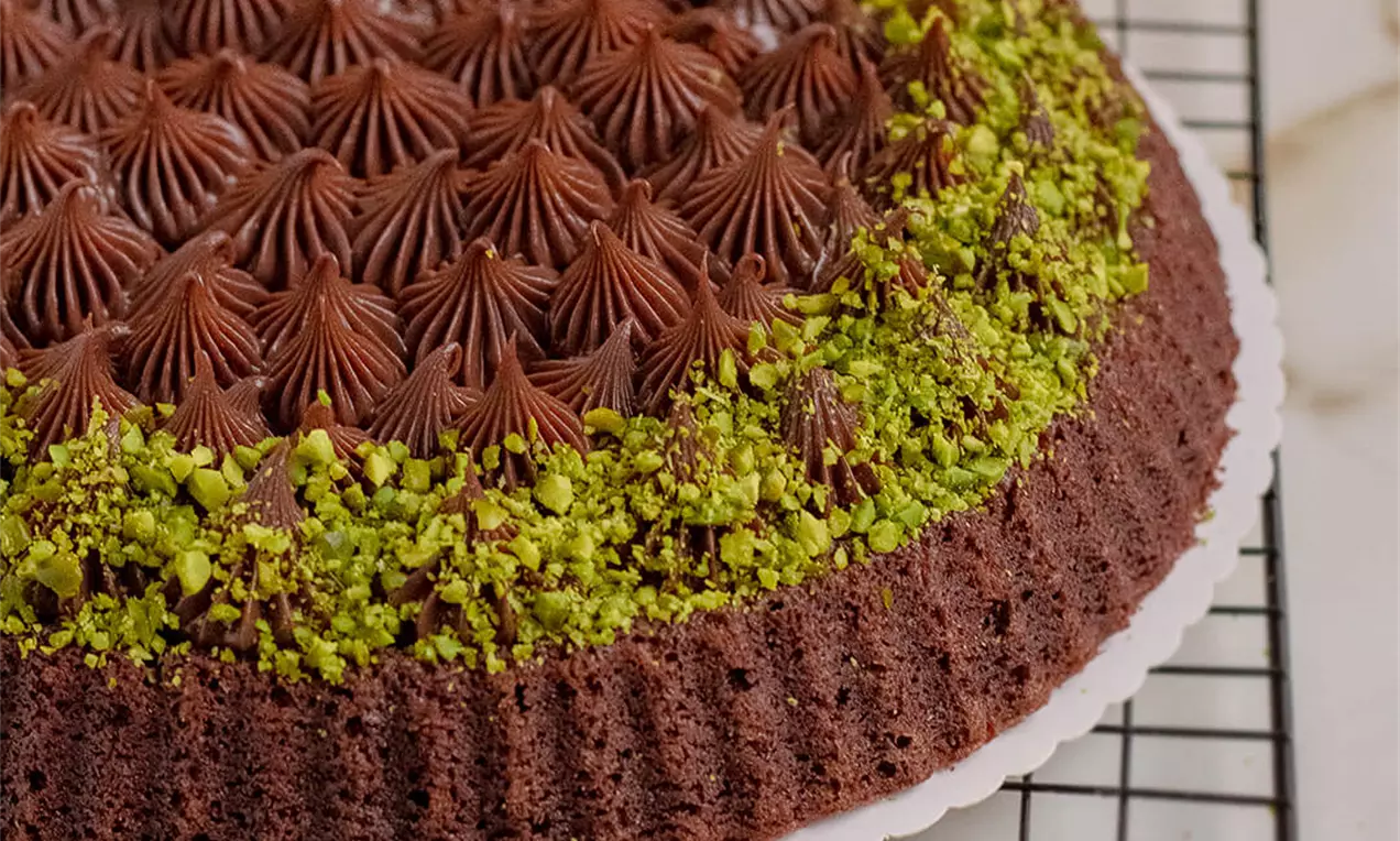 Okrugli čokoladni kolač s bogatim smeđim čokoladnim kremom u obliku rozeta i rubom od sitno sjeckanih zelenih pistacija na bijelom tanjiru