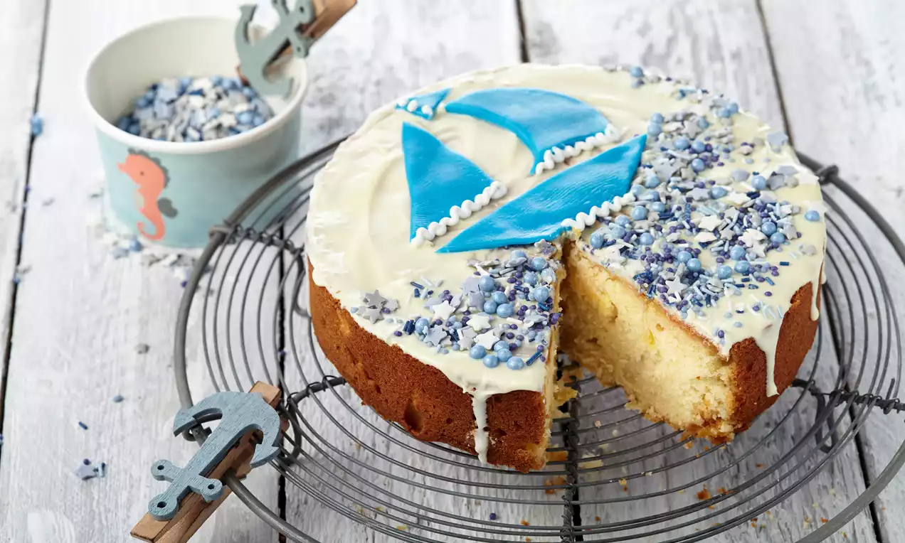 Segelschiffkuchen mit goldbraunem Rührkuchen, Ananas und Marzipan, bedeckt mit weißer Schokolade und kleinem buntem Fondantsegel