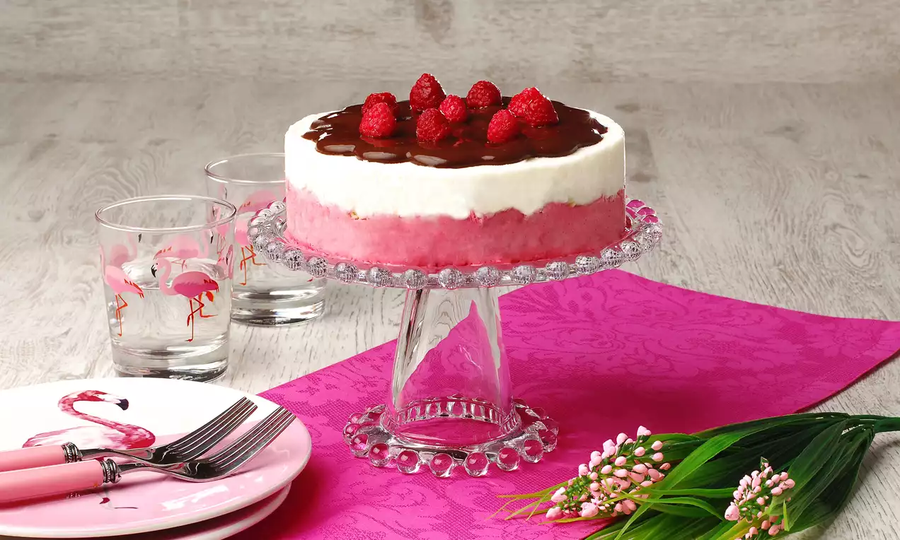 Ahududu ve çikolata sosuyla süslenmiş, pembe-beyaz katmanlı yuvarlak pasta cam ayaklı servis tabağında sergilenmiş
