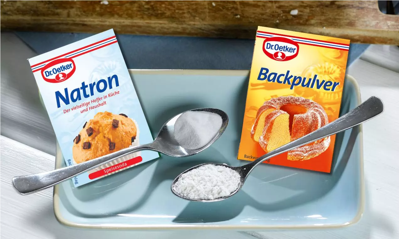 Zwei schräg platzierte Löffel mit weißem Pulver vor bunten Verpackungen für Natron und Backpulver auf hellblauem Porzellanteller
