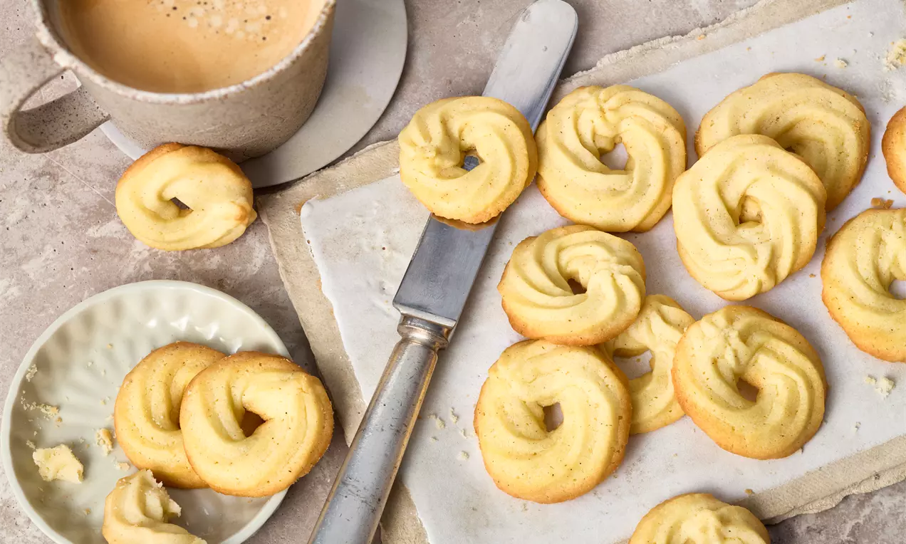 Biscuiți spirală aurii, crocanți pe margini și sfărâmicioși, așezați pe hârtie albă lângă un cuțit și o ceașcă de cafea crem-pe-farfurie
