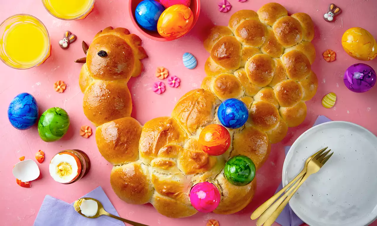 Goudbruine broodhaan met glanzende, gevlochten textuur, versierd met felgekleurde paaseieren op een roze ondergrond met servies