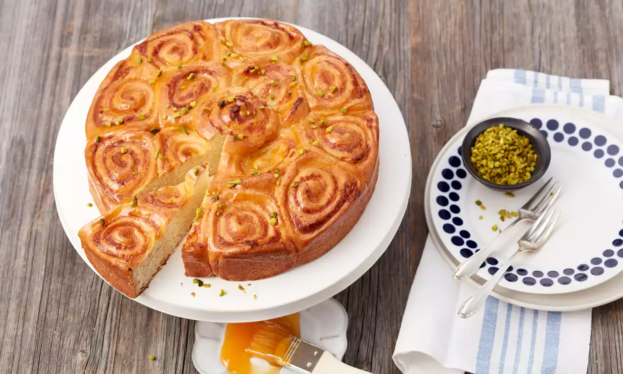 Auf einer weißen Kuchenplatte spiralförmig gebackener Mandel-Rosenkuchen, goldbraun glänzend, mit Pistazien dekoriert und einer gestückelten Auslage