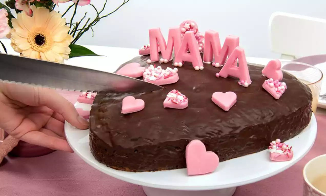 Herzförmiger Kuchen mit glänzender Schokoladenglasur, rosa Zuckerdekorationen und Schriftzug „Mama“ auf weißer Tortenplatte