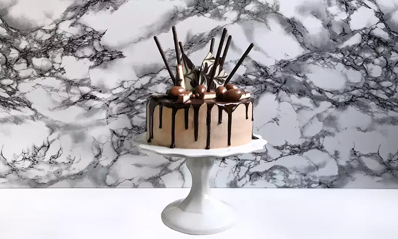 Gâteau cylindrique à la ganache marron clair avec des coulées de chocolat et des décorations en bonbons, bâtonnets et crème sur un présentoir blanc