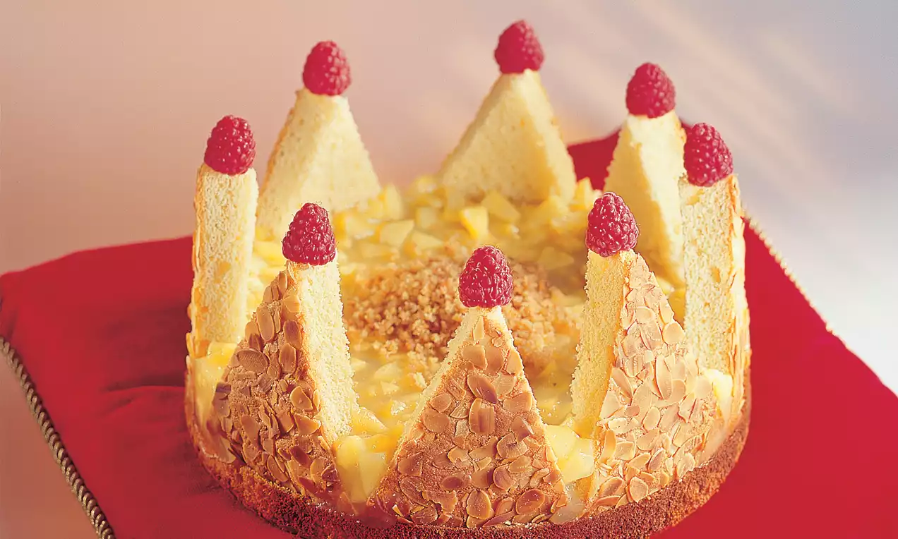 Torta okruglog oblika s kremastim središtem, ukrašena trokutima biskvita posutim bademima i svježim malinama na crvenoj podlozi