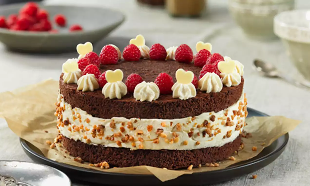 Tarta de capas de bizcocho de chocolate oscuro con relleno de crema suave y almendra picada, decorada con frambuesas y corazones de chocolate blanco sobre un plato
