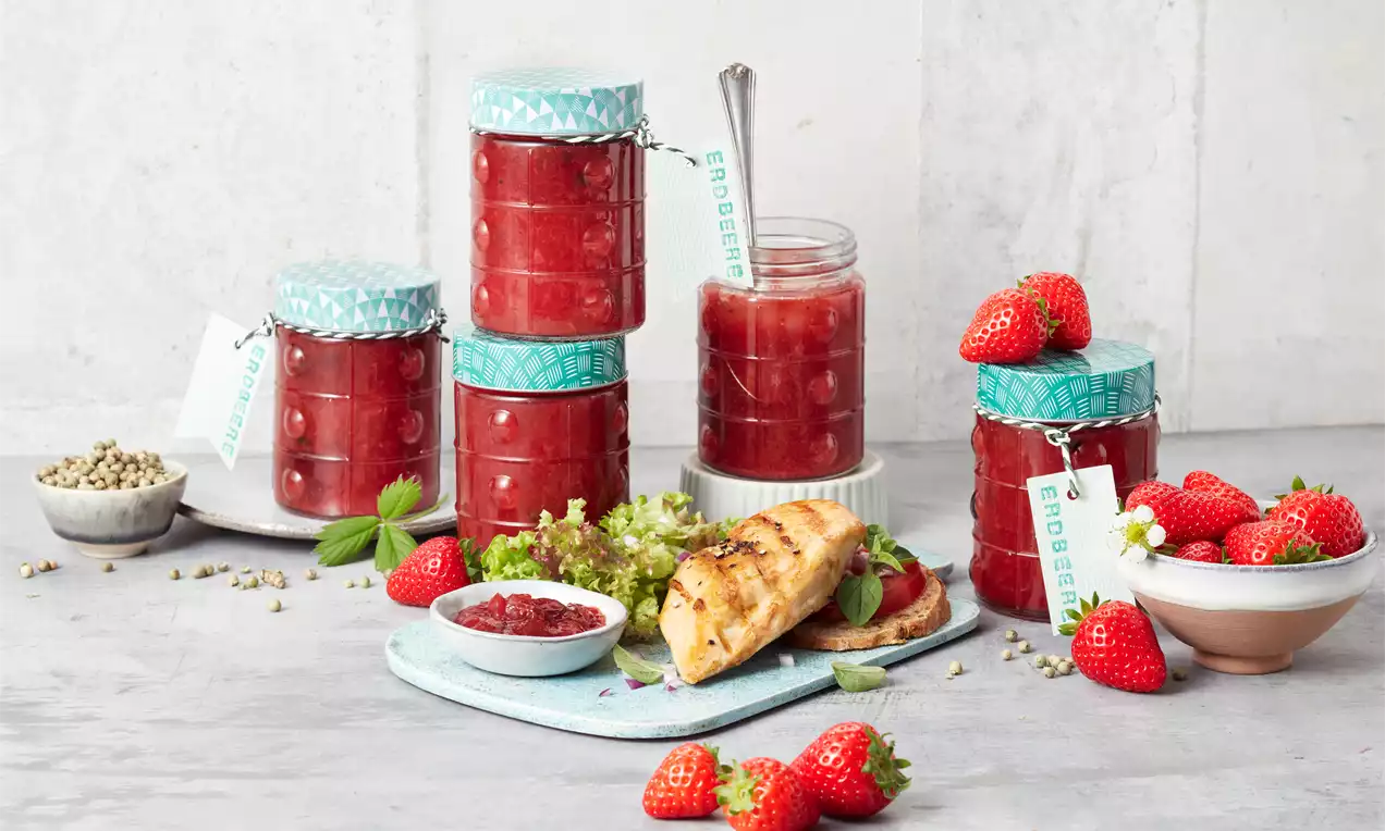 Erdbeer-Chutney in gläsernen Schraubgläsern mit türkisfarbenen Musterdeckeln, frische Erdbeeren, Croissant und Salat auf hellem Untergrund