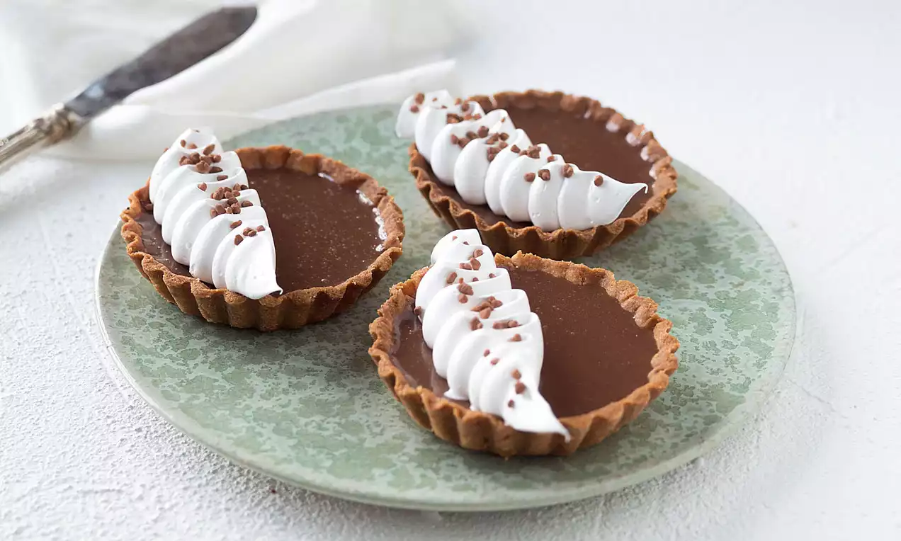 Kolme pientä pyöreää pikkupiirasta kultaruskeilla murokakkupohjilla, täytteenä tumma suklaakerros, valkoista kermavaahtoa ja nonparelleja