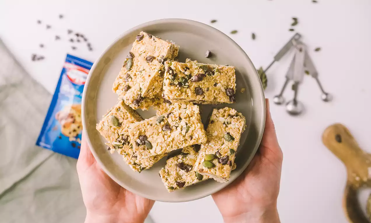 Barras rectangulares de granola dorada con trozos de chocolate y semillas visibles, apiladas en un plato gris sostenido con ambas manos