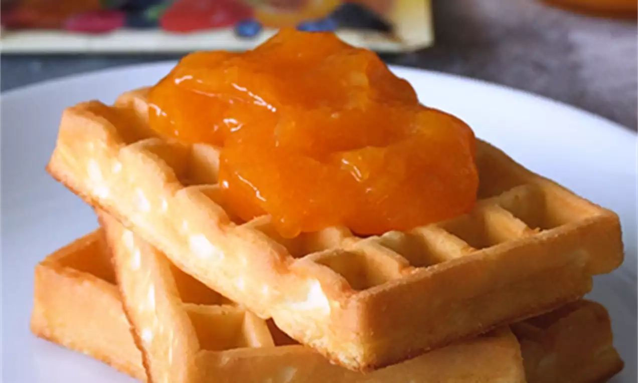 Svetlo oranžna, sijajna marmelada z gladkimi koščki sadja nežno prelita čez hrustljave, zlate vaflje na belem krožniku