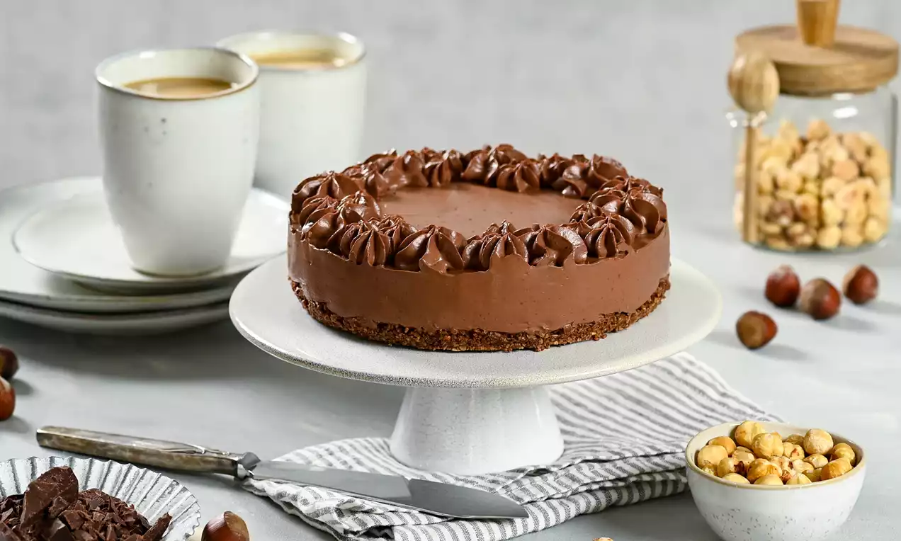 Gâteau rond à la mousse chocolatée brillante sur une base biscuitée avec volutes de crème en bordure, posé sur un plat blanc sur une table garnie de noisettes et accompagnements