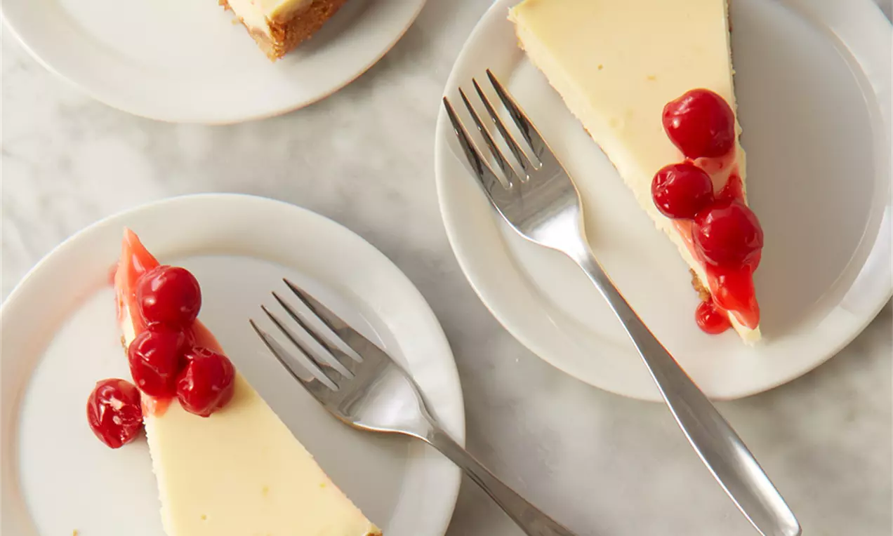 Felie de cheesecake cu blat crocant maro, cremă fină galben-pal și topping de cireșe strălucitoare, pe farfurie albă alături de furculiță argintie