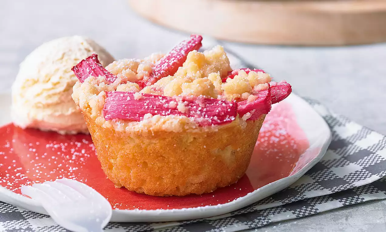Goldbrauner Muffin mit zarten Streuseln und leuchtend roten Rhabarberstücken, leicht mit Puderzucker bestäubt auf rotem Teller mit Eiskugel