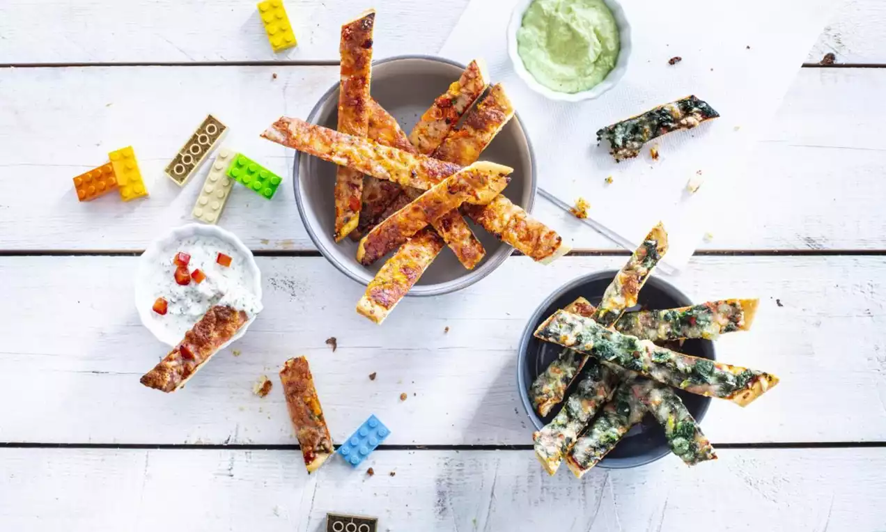 Golden-brown pizza sticks with crispy texture, laid in bowls and scattered on a white wooden surface with dips and colorful Lego bricks