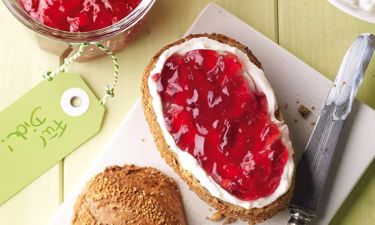 Johannisbeer-Rhabarbergelee in leuchtend roter Farbe auf einer mit Frischkäse bestrichenen Brotscheibe, daneben Messer und Körnerbrötchen