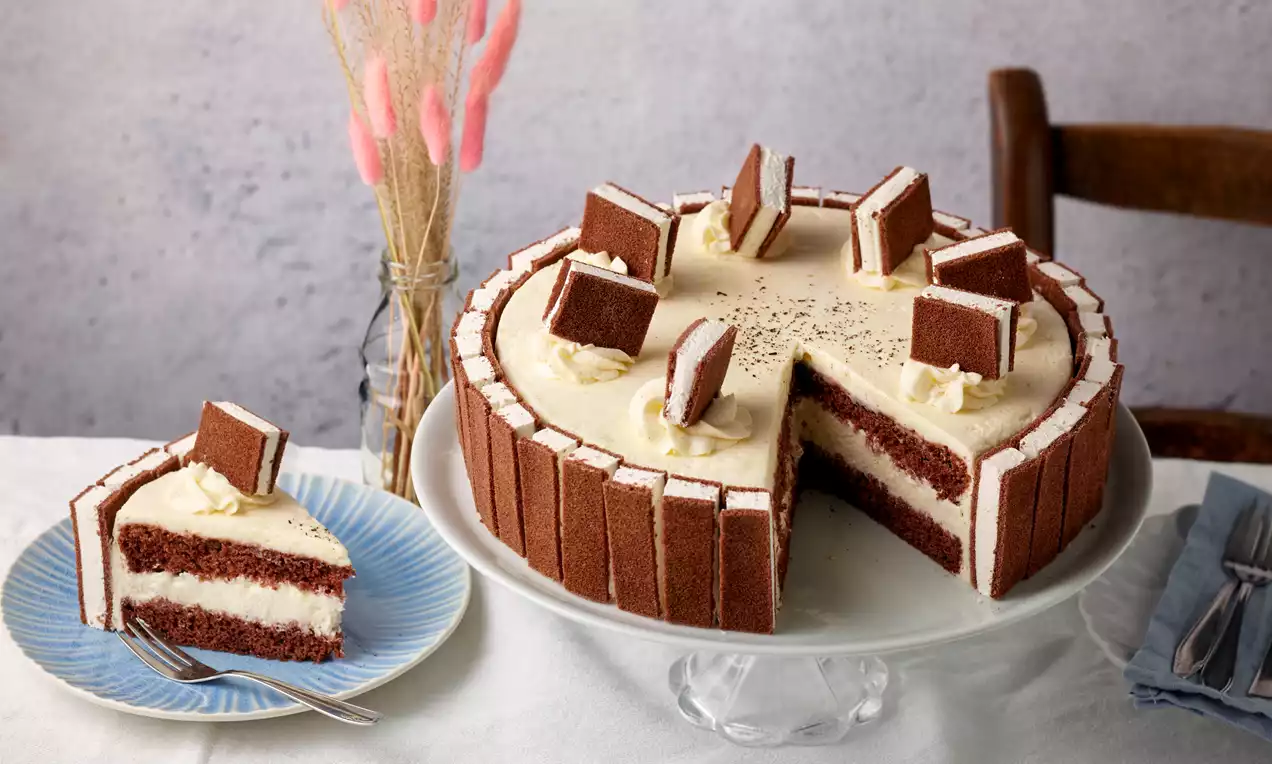 Milchschnitten-Torte mit brauner Umrandung und heller Creme, dekoriert mit Milchschnittenstücken auf einer weißen Tortenplatte