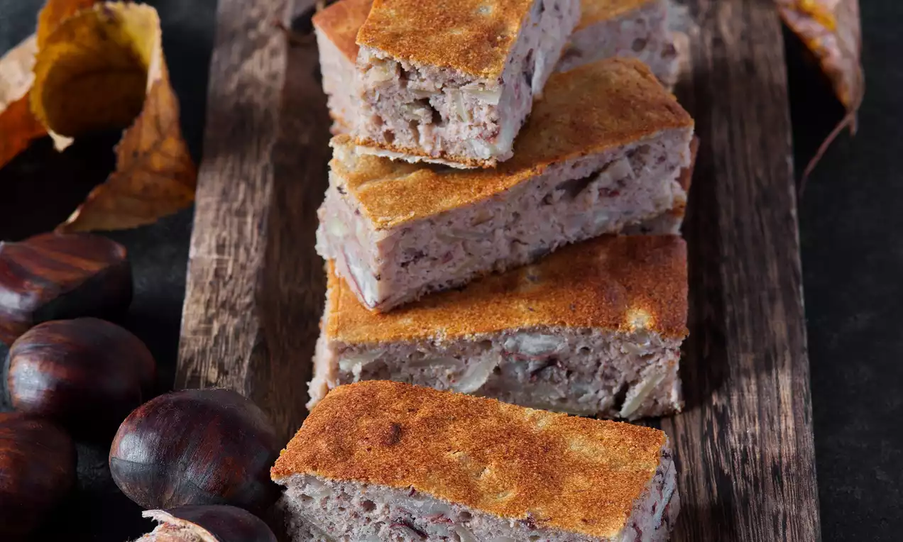 Kaštanové řezy s jemně zlatavou kůrkou, mírně zrnitou texturou, vrstvené do řady na rustikálním dřevěném podnosu obklopeném kaštany a suchými listy