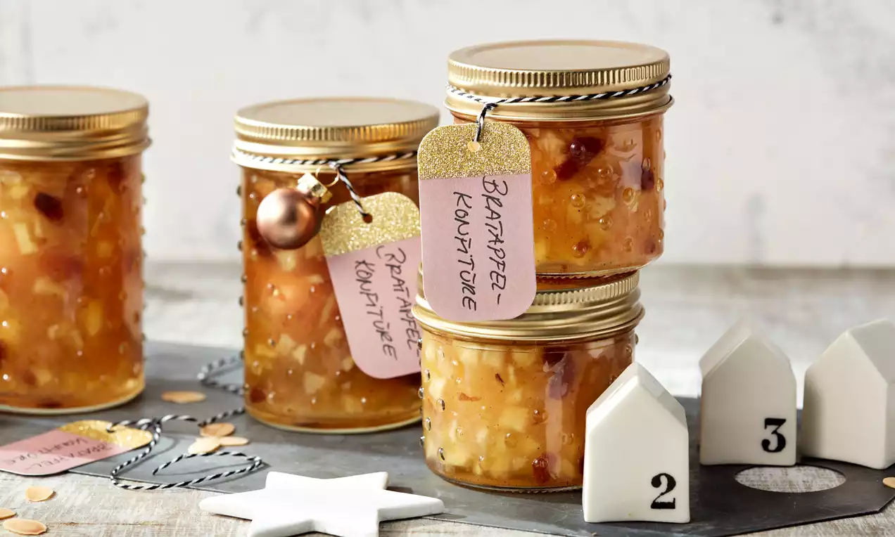 Vier goldbraune Gläser Bratapfel-Konfitüre mit goldenen Deckeln und rosa Anhängern auf dunklem Tisch mit weißen Dekoelementen