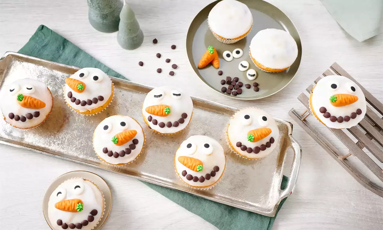 Schneemann-Muffins mit weißem Zuckerguss, orangener Karotten-Deko, Zuckeraugen und Schokoperlen-Lächeln auf silbernem Tablett arrangiert