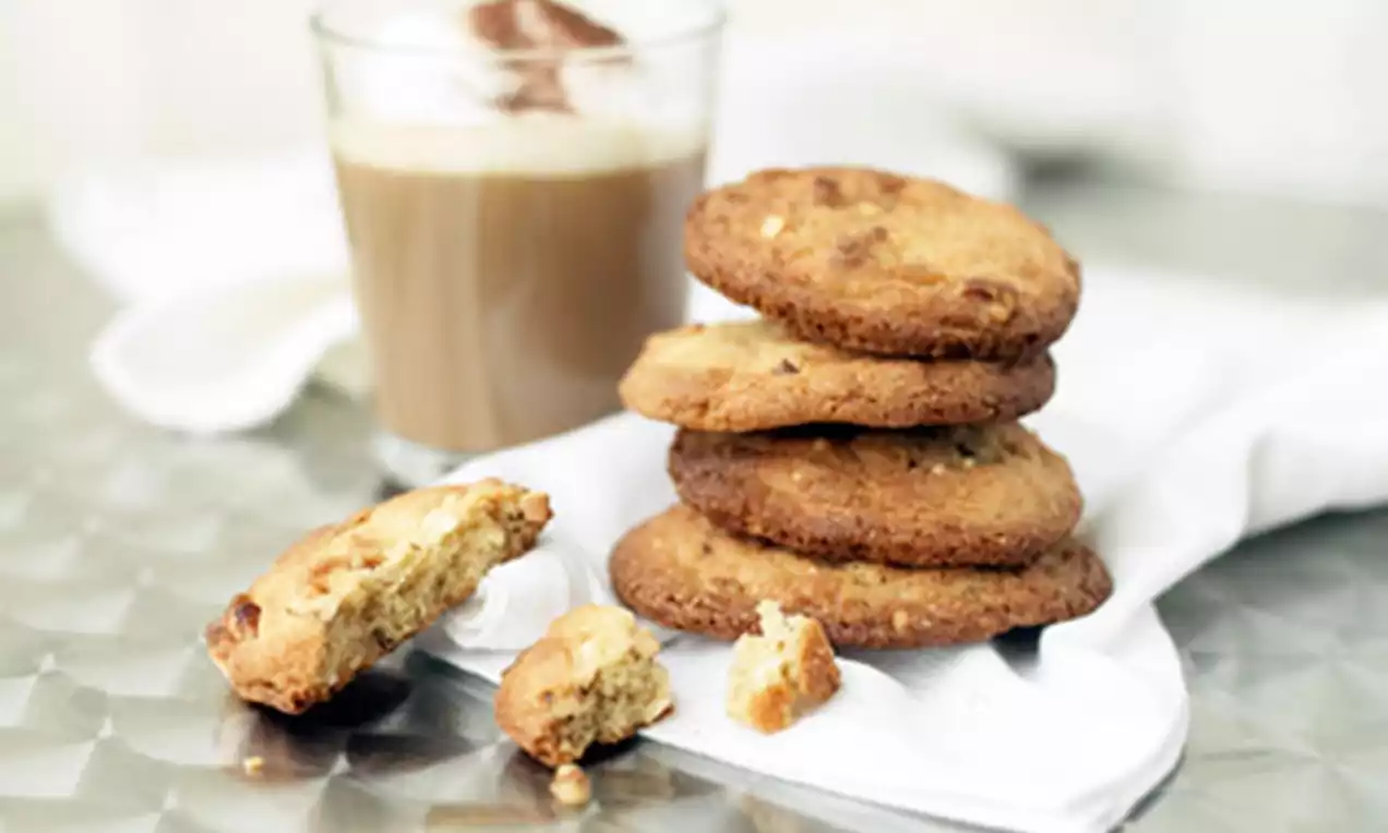Gyldne nøttecookies med sprø kanter og mykere indre stablet på en hvit serviett ved siden av et glass kremet cappuccino