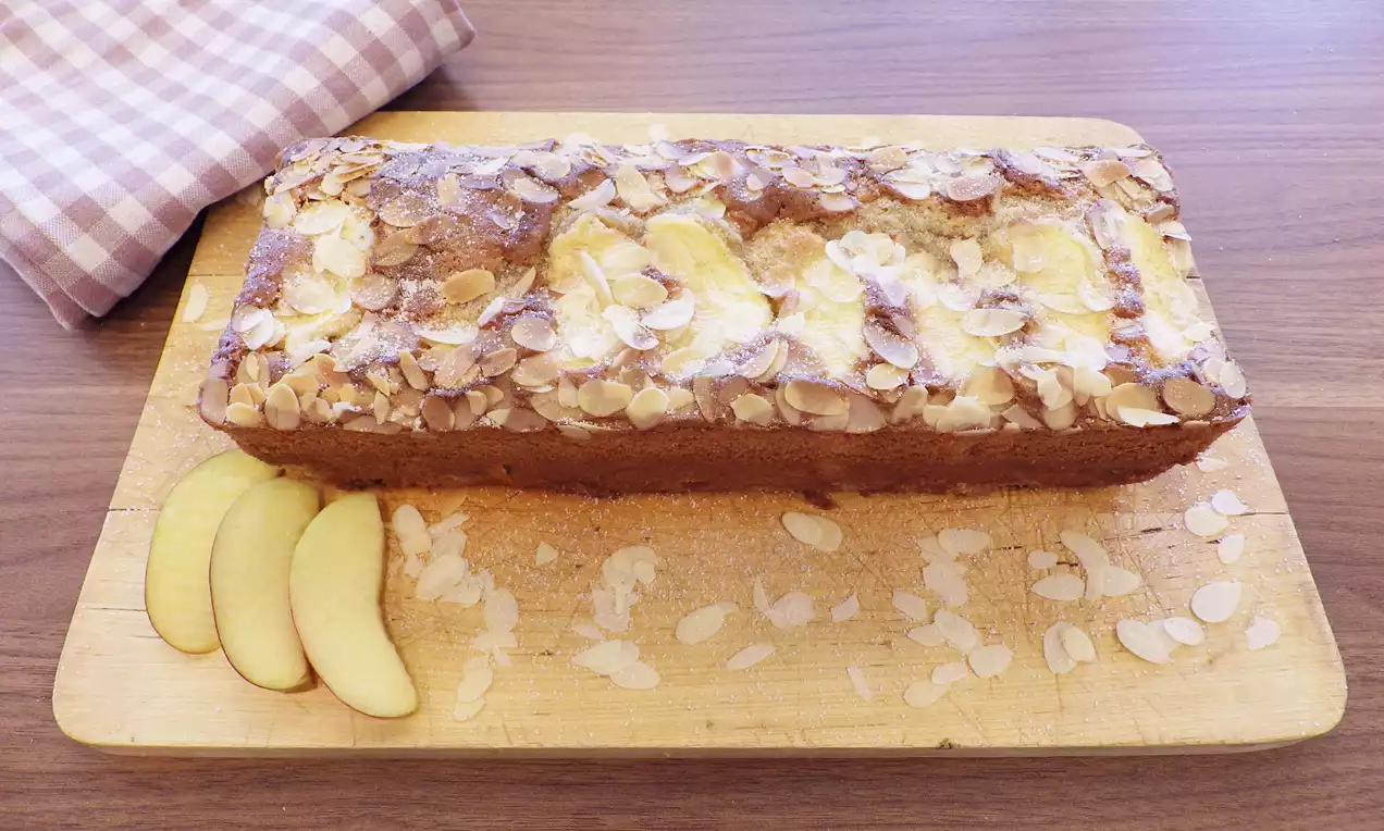 Cake doré rectangulaire sur une planche en bois, garni de tranches de pommes jaunes, éclats d'amandes et entouré de lamelles de pomme