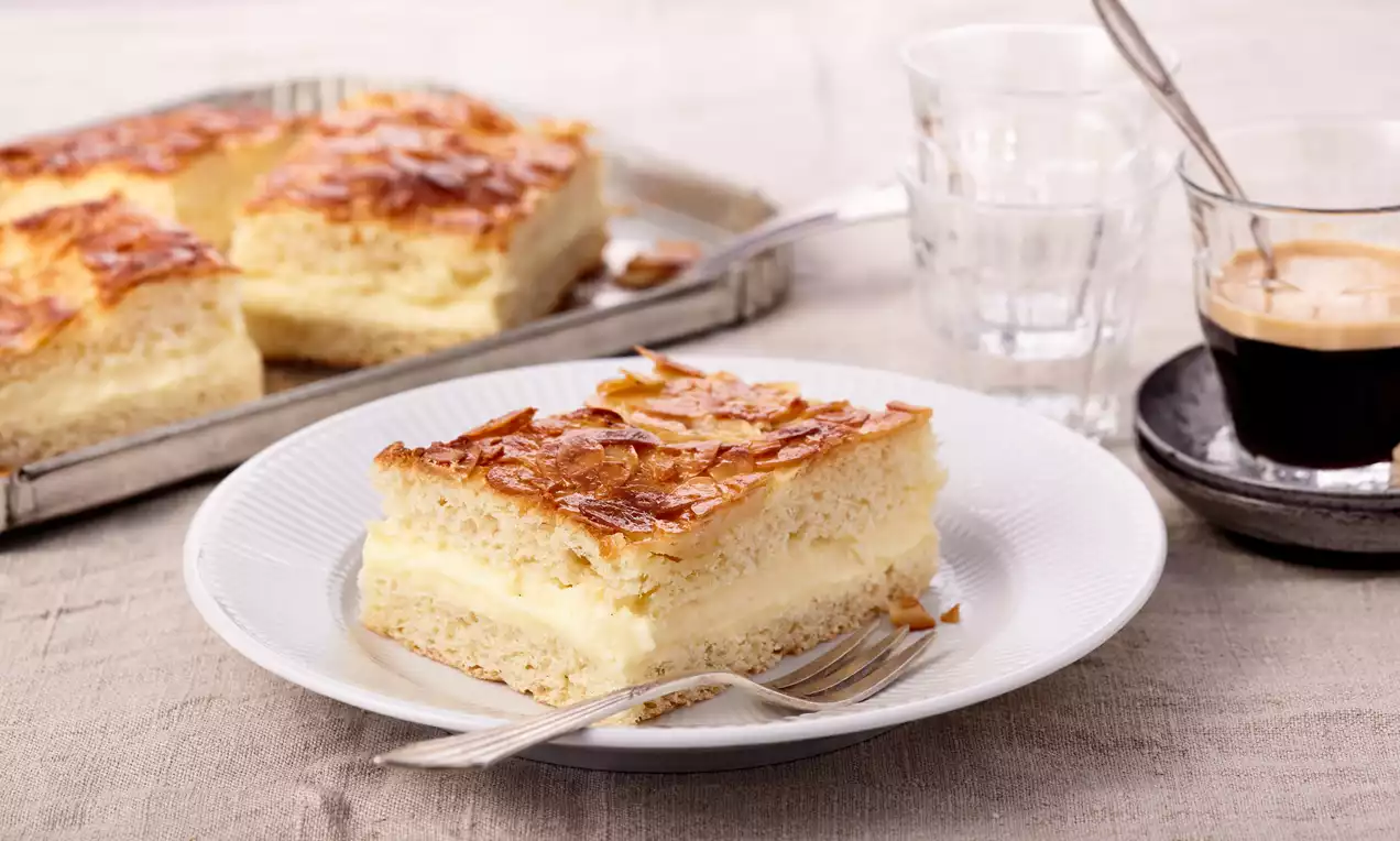 Ein rechteckiges Stück Bienenstich auf weißem Teller mit goldbrauner Mandeldecke, heller Cremefüllung und beige-gelbem Hefeteig, daneben eine Gabel