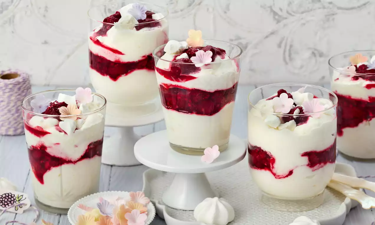 Gläser mit weißer Creme und leuchtend roten Himbeerschichten, dekoriert mit weißen und rosa Blüten aus Zucker auf einem hellen Tablett