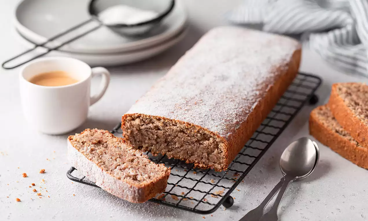 Cake moelleux aux marrons, doré et saupoudré de sucre glace, présenté sur une grille avec quelques tranches coupées et une tasse de café
