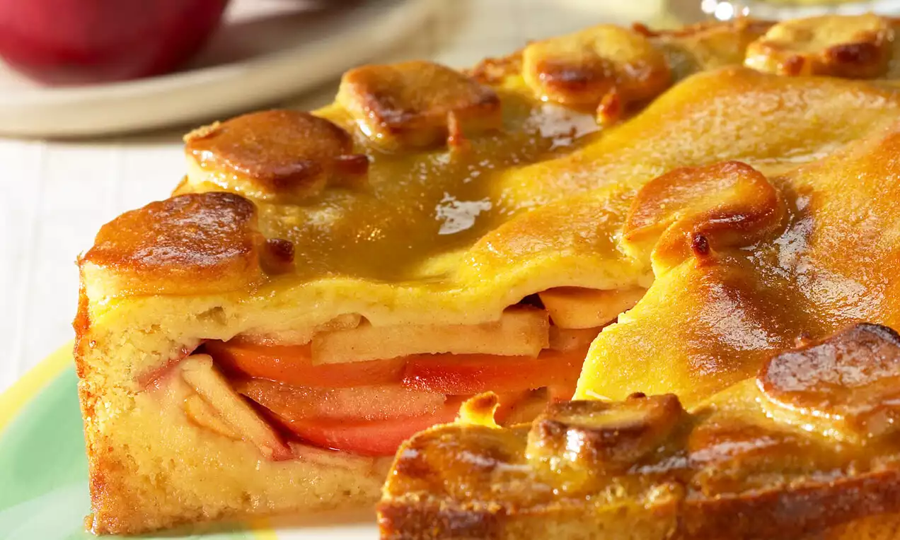 Prăjitură cu blat pufos auriu și umplutură stratificată de felii subțiri de mere, glazurată cu un strat lucios de miere pe o farfurie