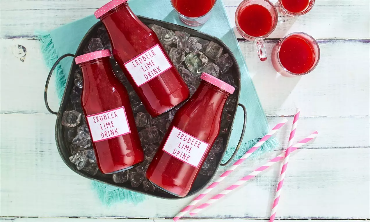 Яркочервени напитки в стъклени бутилки със светлорозови капачки, поставени върху лед в черна тава, с розови сламки и чаши на бял фон