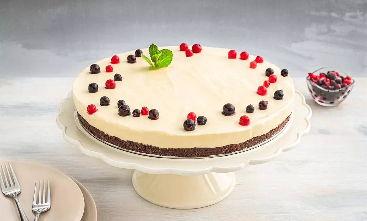 Чийзкейк с гладък крем в бледо жълт цвят върху тъмен бисквитен блат, украсен с червени и черни плодове и свежо ментово листо